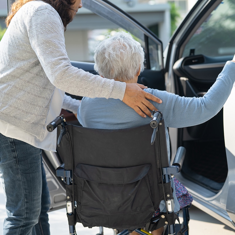 Aide personne âgée : Transfert fauteuil roulant à la voiture. Aidante soutenant une personne âgée en fauteuil roulant pour l'aider à transférer vers une voiture. Scène d'assistance et de mobilité.
