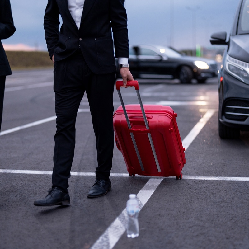 Voyage d'affaires : l'homme en costume et sa valise rouge. Homme d'affaires en costume noir tirant une valise rouge à roulettes dans un parking, près d'une voiture de transfert noire.